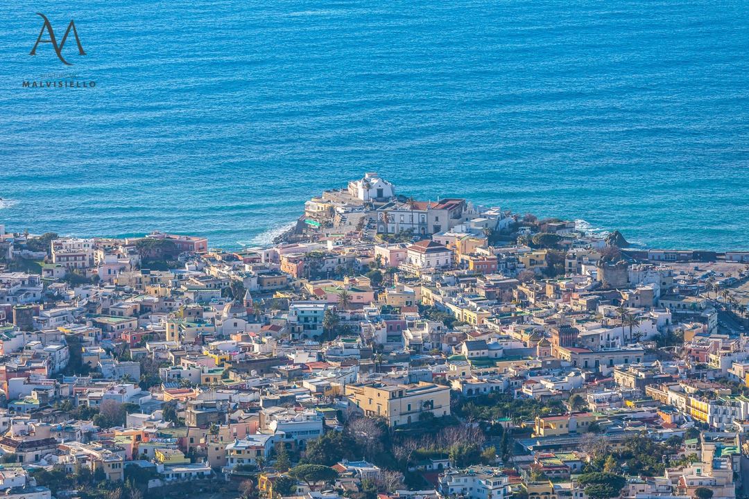 Aerial view of a colorful coastal hillside village overlooking the turquoise Mediterranean Sea with a fortified structure on the waterfront.
